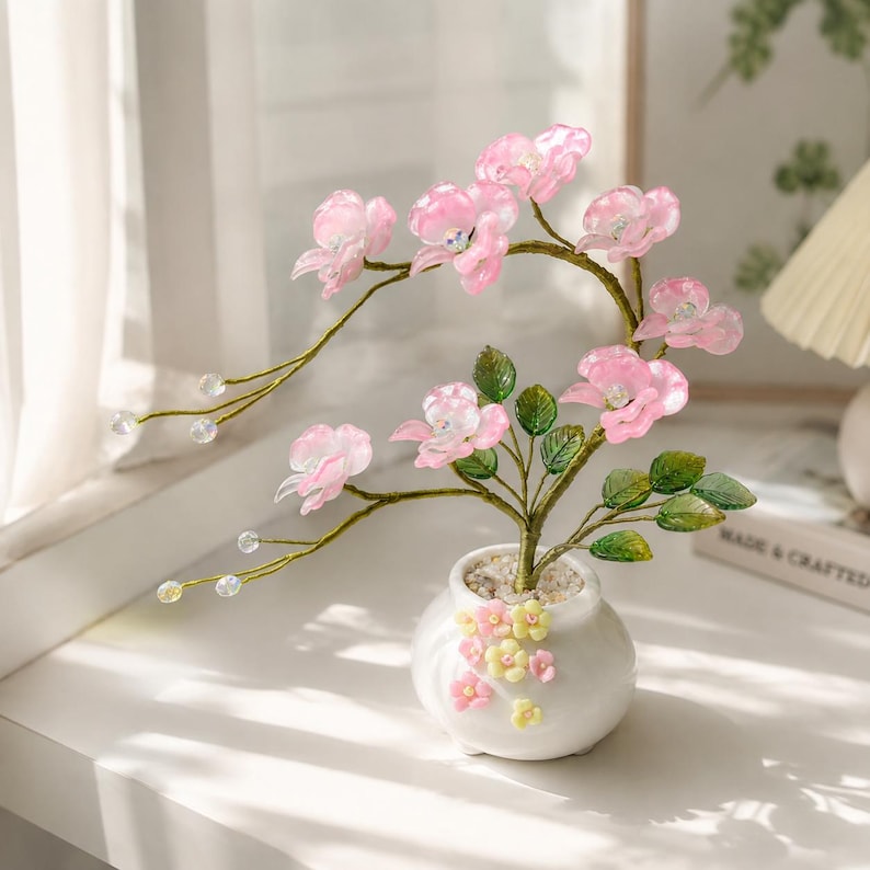 May include: A decorative bonsai tree featuring translucent pink flower blossoms with iridescent centers, green leaves, and thin gold wire branches. The tree is planted in a small white ceramic pot adorned with tiny pink and yellow flower accents.