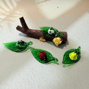 May include: A collection of glass figurines featuring a brown log, green leaves, and various insects. The insects include a ladybug, a yellow bug, and a black and white bug. The leaves are a vibrant green colour.