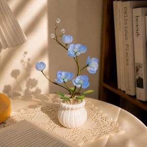 Peut inclure: Une plante artificielle décorative composée de pétales de fleurs bleu clair translucides et de petites perles transparentes sur des tiges en fil métallique fin et flexible. La plante est dans un petit vase en céramique blanc à côtes verticales.