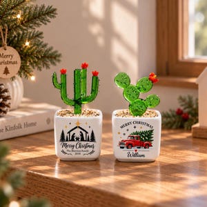 Peut inclure: Deux pots carrés en céramique blanche contenant des cactus en verre vert avec des fleurs rouges. Le pot de gauche présente une scène de nativité noire avec le texte Merry Christmas, The Martinez Family, 2026. Le pot de droite montre un camion rouge avec le texte Merry Christmas, William.