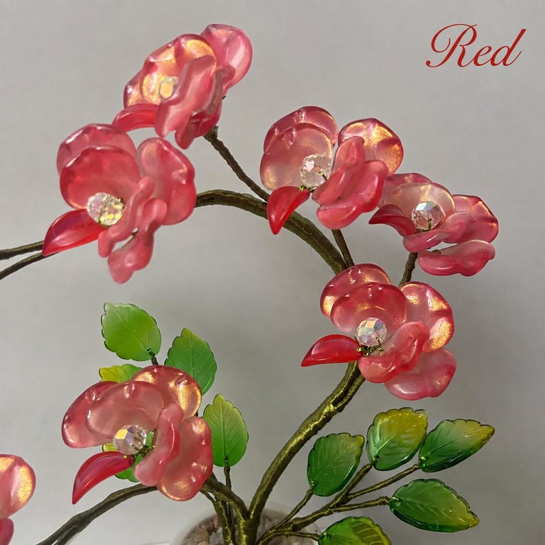 May include: A decorative tree sculpture featuring translucent red flower blossoms with iridescent centers and green leaves attached to thin, dark, wire-like branches. The word Red is printed in a cursive script in the upper right corner.