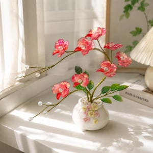 May include: A decorative artificial plant featuring translucent pink flowers with clear bead centers on thin, flexible green stems. The plant is potted in a small, round white ceramic vase adorned with tiny yellow and pink floral accents.