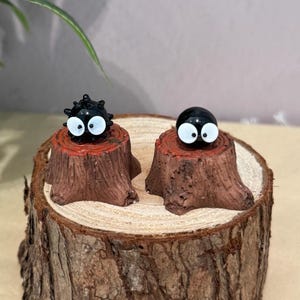May include: Two small, black, round figurines with large white eyes, resembling cartoon characters, sit atop miniature brown tree stumps. The stumps are placed on a large, round, wooden slice, creating a whimsical display.