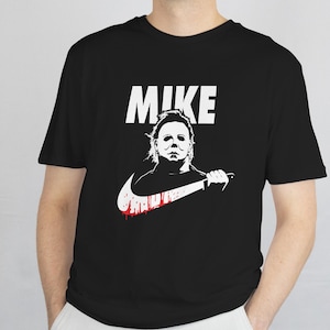 May include: Black t-shirt featuring a white graphic of Michael Myers with a knife, and the word "MIKE" above. The Nike swoosh is part of the design, with a red blood-like splatter.