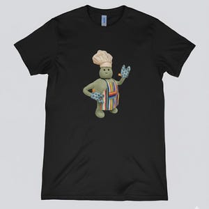 May include: Black t-shirt with a cartoon chef character. The chef is green, wearing a white chef's hat, a colourful striped apron, and blue oven mitts. The character is smiling and waving. The shirt is a Gildan brand.