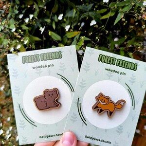 Könnte beinhalten: Zwei Holzpins mit Waldbildern. Ein Pin zeigt einen braunen Bären, der andere einen orangefarbenen Fuchs. Jeder Pin befindet sich auf einer hellgrünen Karte mit dem Text "FOREST FRIENDS" und "wooden pin". Die Karten werden vor einem Hintergrund aus grünem Laub gehalten.