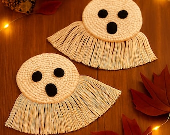 Puede incluir: Dos posavasos hechos a mano con forma de fantasma, con ojos y boca negros, hechos de hilo color crema con flecos. Los posavasos están sobre una superficie de madera, decorados con hojas de otoño, una pequeña calabaza y una vela.
