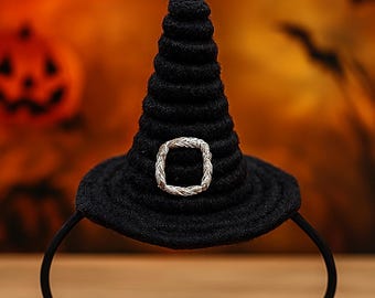 Puede incluir: Una diadema de sombrero de bruja negro con una hebilla plateada. El sombrero está hecho de una tela texturizada y tiene forma de cono. El fondo presenta un tema de Halloween con murciélagos y una calabaza.