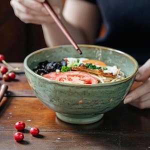 Puede incluir: Un cuenco de cerámica verde lleno de fideos, un huevo frito, rodajas de tomate y hongos negros. Una persona sostiene el cuenco, con palillos chinos listos para comer. Bayas rojas esparcidas sobre la mesa de madera.