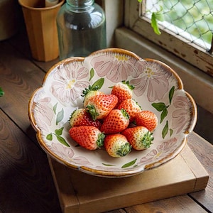 Puede incluir: Un cuenco de cerámica con un diseño floral contiene un montón de fresas frescas. El cuenco tiene un borde festoneado y un borde dorado. Las fresas son rojas con copas verdes. El cuenco está sobre una superficie de madera.