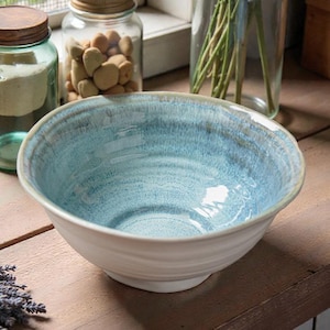 Könnte beinhalten: Eine große, cremefarbene Keramikschüssel mit strukturiertem, hellblauem Innenraum und leicht geriffeltem Rand. Die Schüssel steht auf einer Holzoberfläche, im Hintergrund Gläser und Grün.