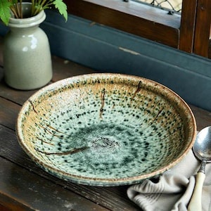 May include: A large, round ceramic bowl with a speckled green and brown glaze. The bowl has a wide rim and a shallow depth. A spoon with a cream-coloured handle rests on a folded cloth next to the bowl. A small vase with green plants is in the background.