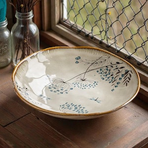 Może przedstawiać: Kremowa ceramiczna miska do serwowania z kwiatowym wzorem w kolorze niebieskim i czarnym. Miska ma szeroki, płytki kształt z brązową obwódką. Miska stoi na drewnianej powierzchni.