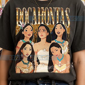 Può includere: T-shirt grigio scuro con la scritta dorata "POCAHONTAS" sopra un collage di sei donne animate. Le donne hanno lunghi capelli neri e indossano abiti diversi. La t-shirt è realizzata in materiale morbido.