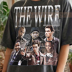 以下が含まれることがあります： メタリックシルバーのフォントで「THE WIRE」と書かれたチャコールグレーの半袖Tシャツ。テキストの下には、白い稲妻のアクセントが施された暗い背景をバックに、テレビ番組の6人のキャラクターがグラフィックプリントされています。