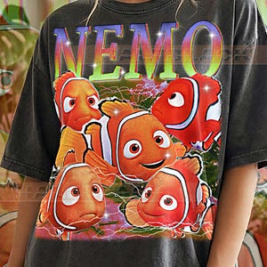 May include: Dark gray t-shirt featuring a colorful graphic of five clownfish with the word "NEMO" in a rainbow font. The text "PEPPER COMFORT COLORS" is printed at the bottom of the shirt.