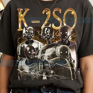 May include: Dark gray t-shirt featuring a graphic of K-2SO droids from the Star Wars franchise. The text "K-2SO" is in gold, with a lightning bolt design. The shirt is worn by a person wearing a gold necklace and blue jeans.