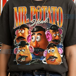 May include: Dark gray t-shirt featuring a graphic of Mr. Potato Head characters with mustaches and hats. The shirt has the text "MR. POTATO Head" in a retro style. The design includes lightning bolts and a vintage aesthetic.