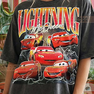May include: Dark gray t-shirt featuring a graphic of Lightning McQueen from the Cars movie. The design includes the text "LIGHTNING McQueen" in a yellow and red font, with multiple images of the character. The shirt is labeled "PEPPER COMFORT COLORS".