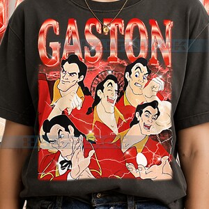 May include: Dark gray t-shirt featuring a graphic of Gaston from Beauty and the Beast. The design includes multiple images of Gaston in a red and yellow outfit, with the name "GASTON" in large red letters above.