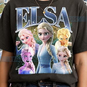 May include: Dark grey t-shirt with the word "ELSA" in a blue, lightning-bolt style font. The shirt features a graphic of Elsa from the film Frozen, with multiple images of her in different poses and outfits.