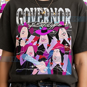 Può includere: T-shirt grigio scuro con una grafica del governatore Ratcliffe del film d'animazione. Il design include la parola "GOVERNOR" in un font metallico sopra l'immagine del personaggio. Il personaggio è raffigurato con un cappello viola e un'espressione severa.