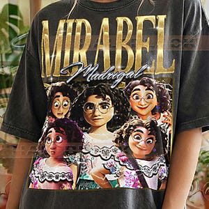 Puede incluir: Camiseta gris oscuro con el nombre "MIRABEL" en letras doradas sobre un gráfico de Mirabel Madrigal de la película Encanto. La camiseta también tiene el texto "Madrigal" en cursiva. Las palabras "PEPPER COMFORT COLORS" están impresas en la parte inferior.