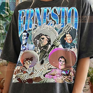 May include: Dark gray t-shirt featuring a graphic of Ernesto de la Cruz with a guitar and other musicians. The text "ERNESTO De la Cruz" is in blue and white. The shirt also has the text "PEPPER COMFORT COLORS".