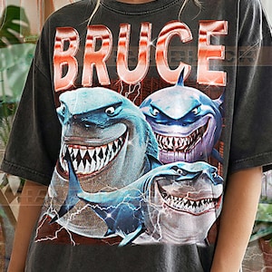 Op de afbeelding: Donkergrijs T-shirt met een afbeelding van drie cartoonhaaien met scherpe tanden en blauwe en witte kleuren. Het woord "BRUCE" is in grote, oranje letters boven de haaien gedrukt. De tekst "PEPPER COMFORT COLORS" staat onderaan.