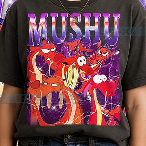 Może przedstawiać: Ciemnoszara koszulka z grafiką postaci Mushu z filmu Mulan. Projekt zawiera napis "MUSHU" w fioletowo-srebrnym gradiencie, z kilkoma czerwonymi smokami na fioletowym tle.