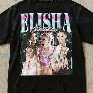May include: Black t-shirt featuring a retro-style graphic with the name "ELISHA Cuthbert" in a colorful, iridescent font. The graphic includes images of several women with lightning bolt accents, creating a vintage aesthetic.