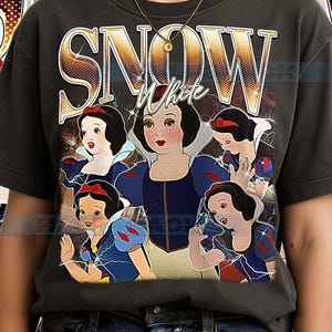 Peut inclure: T-shirt gris foncé avec un graphisme de Blanche-Neige dans différentes poses. Le mot "SNOW" est en lettres dorées au-dessus de "White" en écriture cursive. Blanche-Neige porte une robe bleue et jaune avec un nœud rouge.
