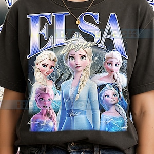 May include: Dark grey t-shirt with the word "ELSA" in blue, and "Frozen" underneath. The shirt features multiple images of Elsa from the animated film, in different poses and outfits. The design has a vintage, distressed appearance.