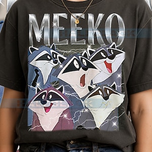 Può includere: T-shirt grigio scuro con la scritta "MEEKO" in lettere argentate. Sotto, una grafica mostra cinque procioni con maschere nere, su uno sfondo di fulmini. Un capo di abbigliamento casual.