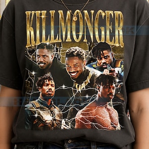 May include: A charcoal gray short-sleeve t-shirt featuring a graphic print of the character Killmonger in various poses. The text KILLMONGER is printed in large gold letters at the top, with white lightning bolt accents surrounding the character images.