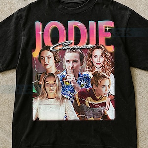 May include: Black t-shirt featuring a retro-style graphic with the name "JODIE" in pink and yellow, with multiple images of a woman. The design has a vintage aesthetic with lightning bolt accents.