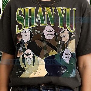 Peut inclure: T-shirt gris foncé avec un graphisme du personnage Shan Yu du film d'animation Mulan. Le design comprend le nom du personnage en vert et jaune, avec plusieurs images de Shan Yu.
