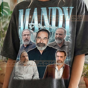 Puede incluir: Camiseta gris oscuro con el nombre "MANDY" en un diseño azul y blanco sobre un fondo de paisaje urbano. La camiseta presenta un gráfico de un hombre con gafas y barba en varias poses. El texto "Pepper Comfort Colors" está en la parte inferior.