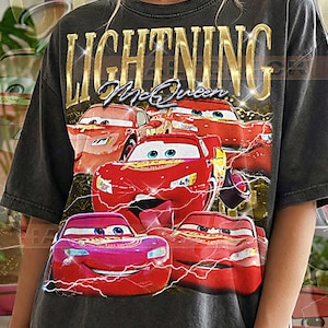 May include: Dark gray t-shirt featuring a graphic of Lightning McQueen from the Cars movie. The design includes multiple red race cars with lightning bolts and the text "LIGHTNING McQueen" in gold lettering. The shirt also has the text "PEPPER COMFORT COLORS".