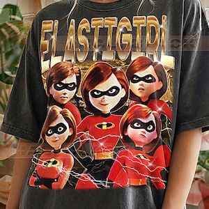 May include: Dark gray t-shirt featuring a graphic of the characters from the animated film, The Incredibles. The shirt has the word "ELASTIGIRL" in gold lettering across the top. The bottom of the shirt reads "PEPPER COMFORT COLORS".