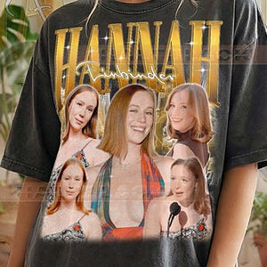 Puede incluir: Camiseta gris oscuro con un gráfico dorado que dice "HANNAH" encima de "Finbinder". La camiseta presenta un collage de imágenes de una mujer. El texto "PEPPER COMFORT COLORS" está impreso en la parte inferior.
