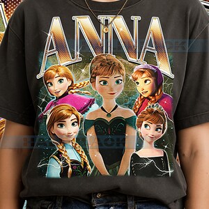 May include: Dark grey t-shirt with the word "ANNA" in a gold, retro font above a graphic of the animated character Anna from the film Frozen. The graphic shows Anna in various poses with a teal and blue background.