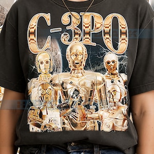 May include: Dark gray t-shirt featuring a graphic of C-3PO and other golden robots from Star Wars. The text "C-3PO" is in a gold, textured font above the robots. The design includes a space-themed background.