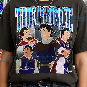 Peut inclure: T-shirt gris foncé avec un imprimé représentant un prince de dessin animé et le texte "THE PRINCE" en bleu et blanc. Le motif comprend plusieurs illustrations du prince dans différentes poses, sur fond de ciel étoilé.