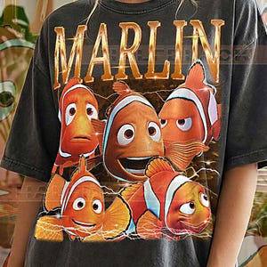 May include: Dark gray t-shirt featuring a graphic of several cartoon clownfish with the word "MARLIN" in gold lettering. The shirt has the text "PEPPER COMFORT COLORS" at the bottom.
