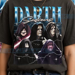 May include: Dark gray t-shirt featuring a graphic of Darth Sidious and other characters. The word "DARTH" is in large, blue, stylized text above the image, with "Sidious" in a cursive font. The shirt has a vintage, distressed look.