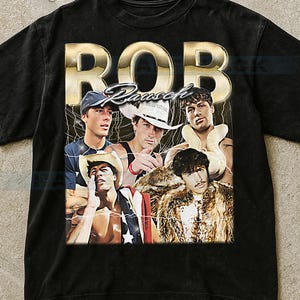 Puede incluir: Camiseta negra con un diseño gráfico de estilo vintage. La palabra "ROB" en letras doradas sobre un collage de cinco hombres. El collage incluye imágenes de los hombres con varios atuendos, incluyendo sombreros de vaquero y pieles.