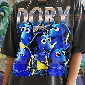 May include: Dark gray t-shirt featuring a graphic of the character Dory from the movie "Finding Nemo". The word "DORY" is printed across the top in a silver, metallic font. The graphic includes multiple blue fish with yellow fins.