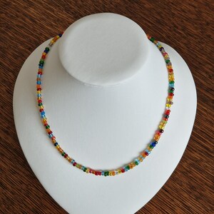 Op de afbeelding: Een enkelstrengs ketting bestaande uit kleine, ronde, veelkleurige glaskralen. De kralen hebben een verscheidenheid aan levendige tinten, waaronder rood, oranje, geel, groen, blauw en transparante tinten, gerangschikt in een herhalend patroon.