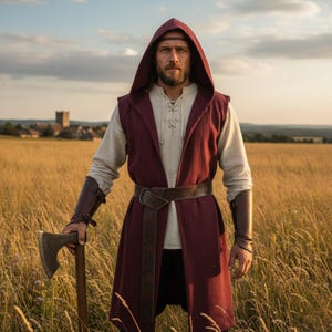 May include: A burgundy hooded vest and matching arm bracers are worn over a cream-colored tunic. A brown leather belt and headband complete the medieval-inspired outfit. The person is holding a wooden-handled axe in a field of tall grass.
